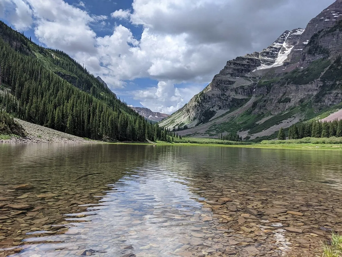 Clear mountain lake over smooth stones, with a pine slope and snow-streaked rock peaks under summer clouds