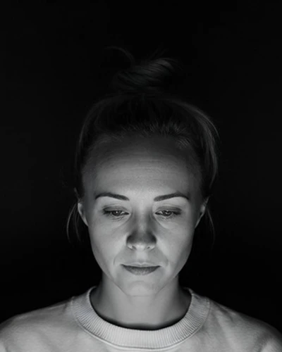 Black-and-white portrait of Katie Rutten, hair tied up, in a light sweater, her face lit by a laptop screen in a dark room