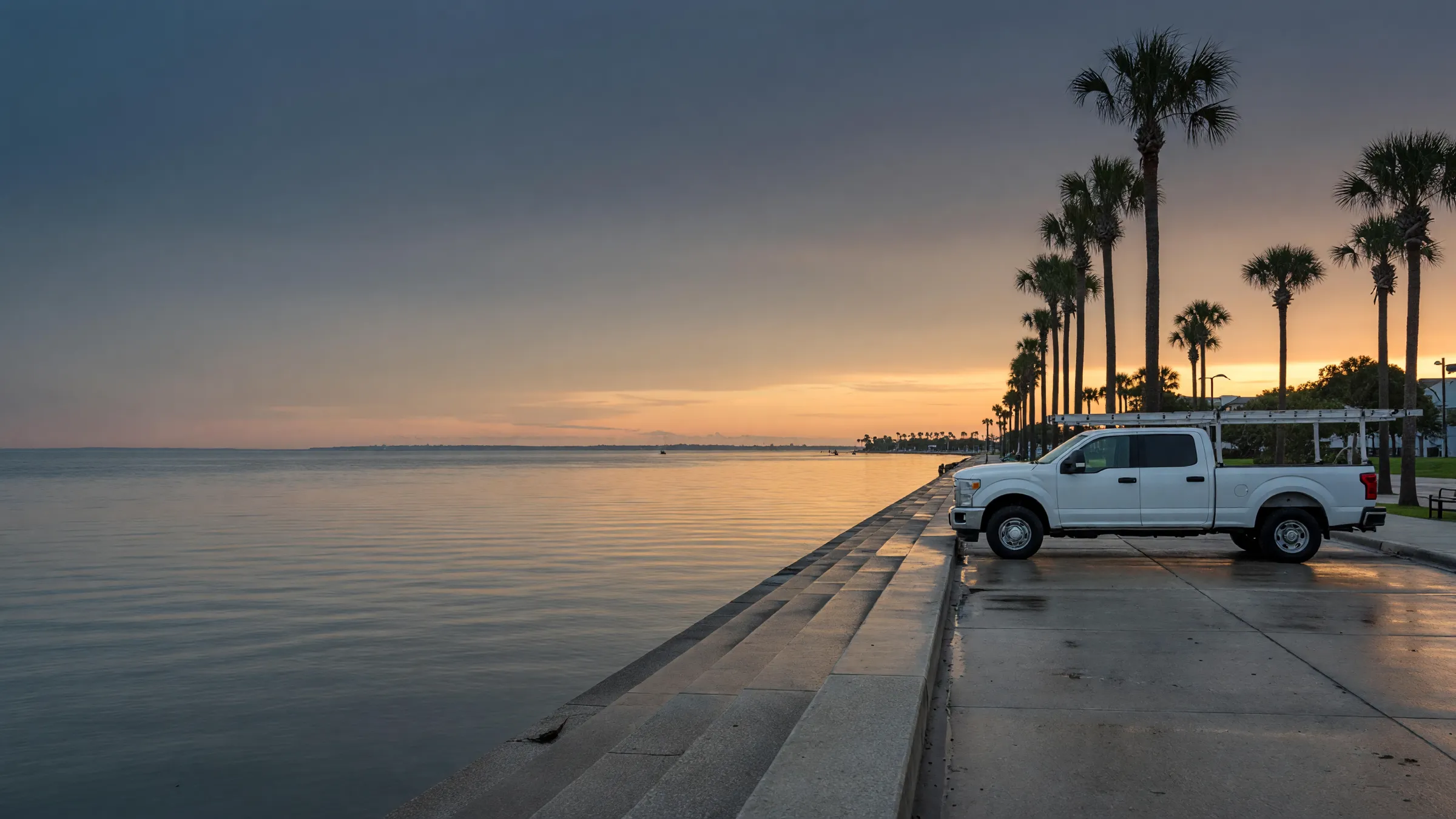 A white work pickup parked under palm trees by the Corpus Christi bayfront seawall at sunrise.