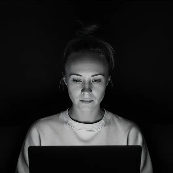 Katie Rutten working on a laptop, lit by its screen, black and white portrait.