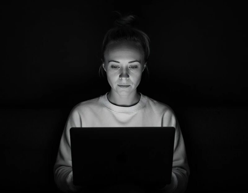 Black-and-white photo of Katie Rutten, hair in a bun, in a light sweater, working on a laptop in a dark room