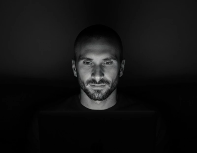 Black-and-white photo of Kyle Rutten, a bearded man with a shaved head, lit by a laptop's glow in a dark room