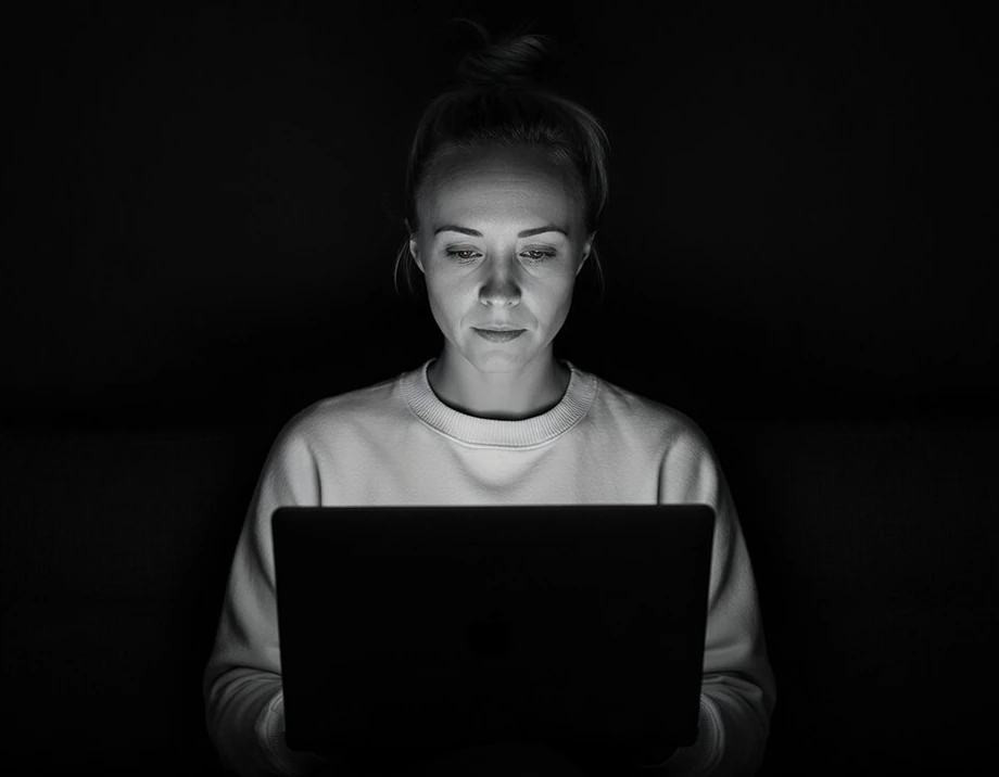 Katie Rutten in a light sweater, working on a laptop in a dark room, in black and white
