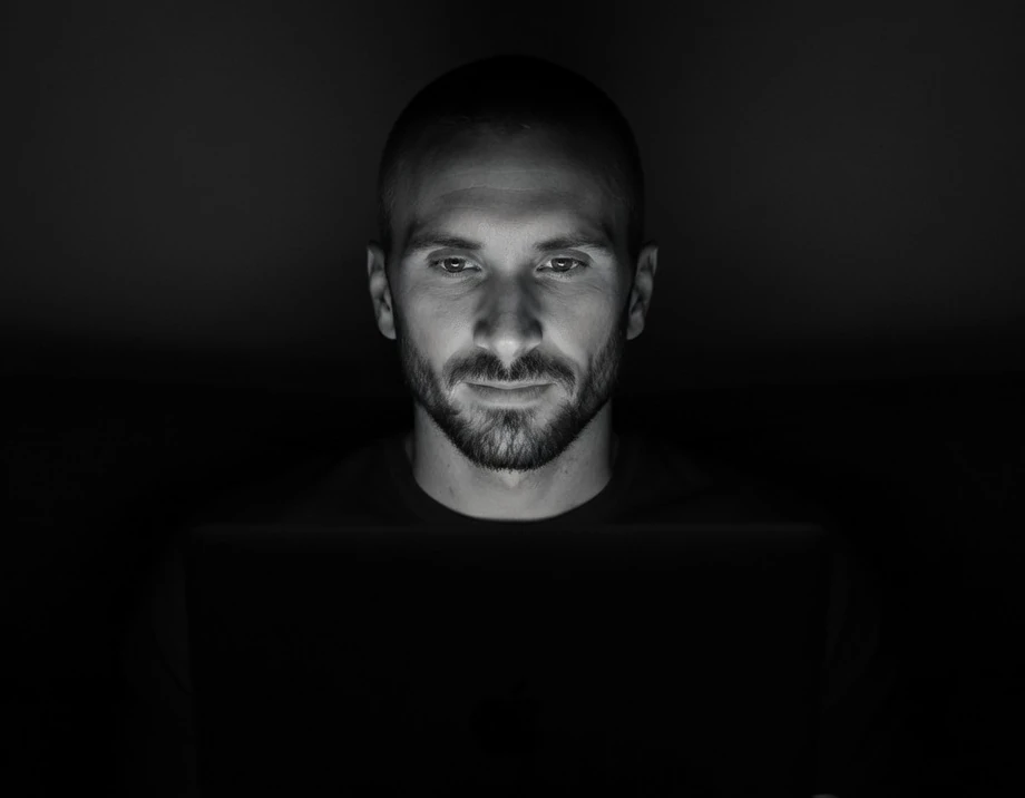 Kyle Rutten, a bearded man with close-cropped hair, lit by a laptop screen in a dark room, in black and white