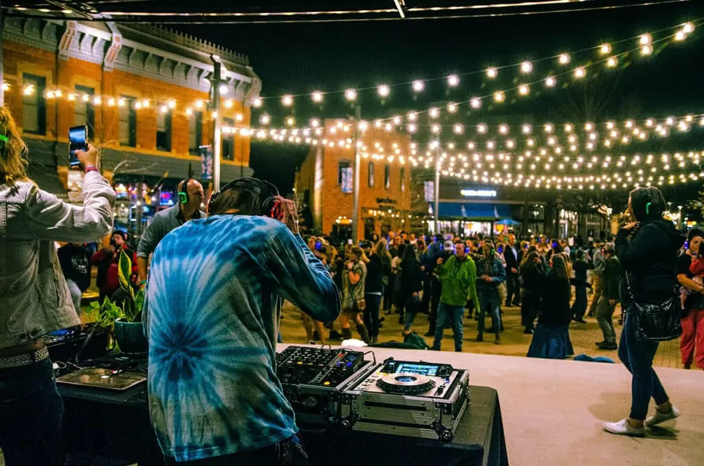 Old Town Square, Fort Collins, at night: string lights, brick storefronts, a crowd in light-up headphones, a DJ at the decks