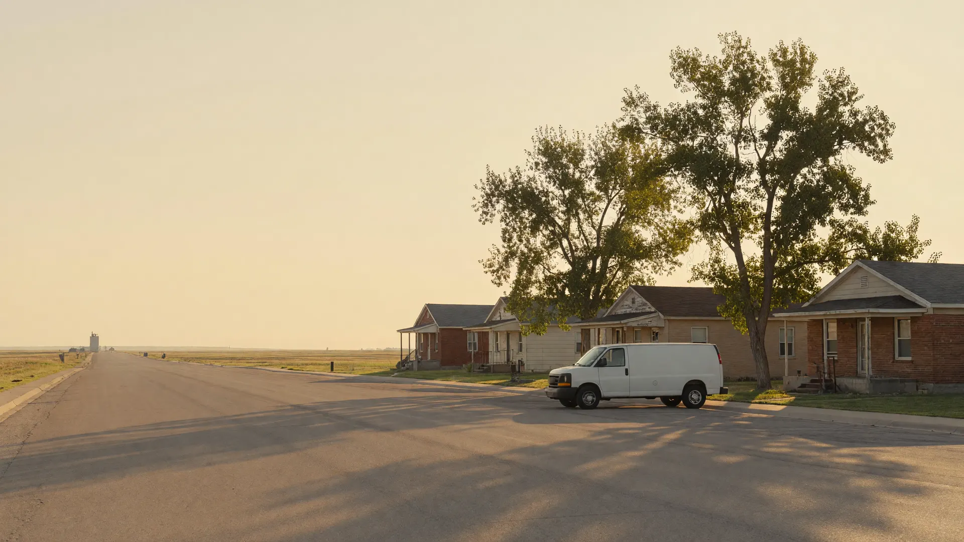 A white service van at the curb on a quiet Greeley street of brick houses and cottonwoods, open plains beyond