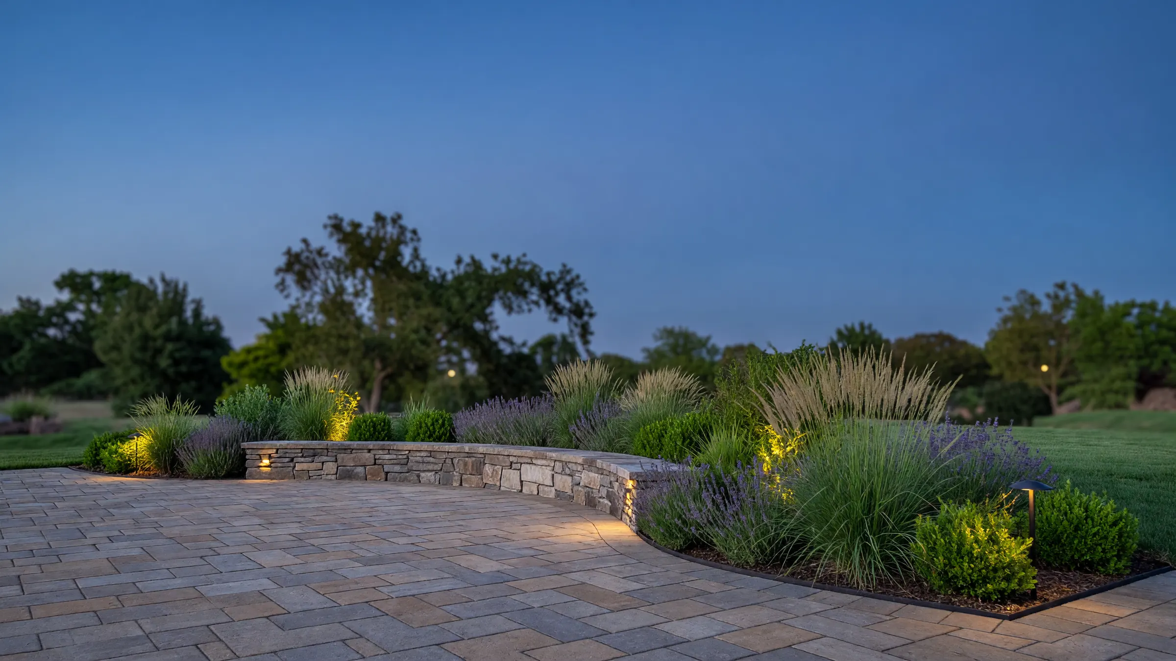 Backyard at dusk with a paver patio, a curved stone seat wall and planting beds, landscape lights just switched on