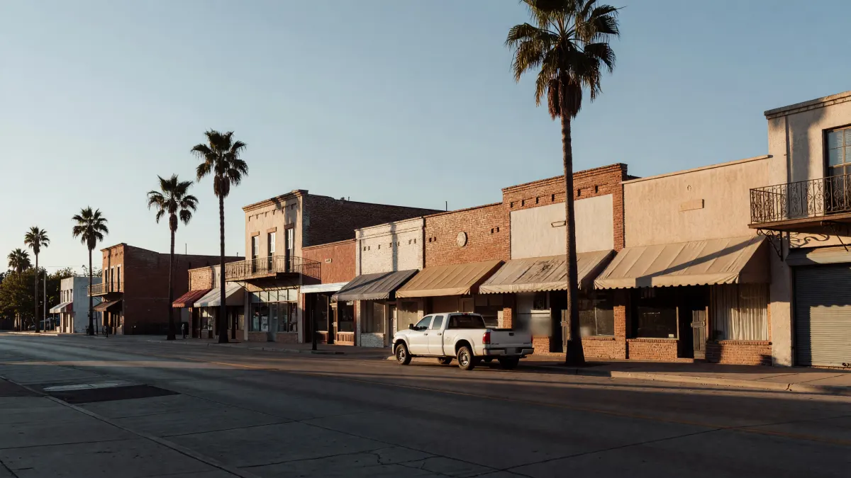Low brick and stucco storefronts with awnings and palm trees on a quiet street, a white pickup at the curb