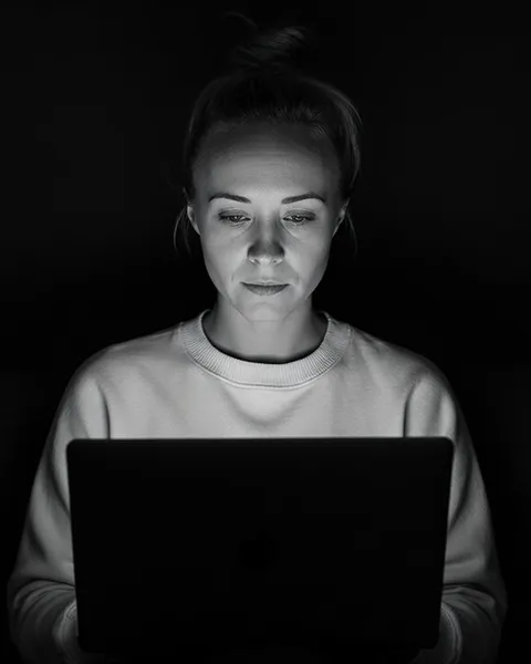 Katie, her hair in a bun, wearing a light sweater and working on a laptop in a dark room