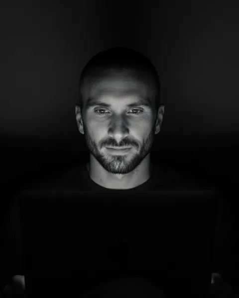 Kyle, a bearded man with a shaved head, lit by the glow of the laptop he's working on in a dark room