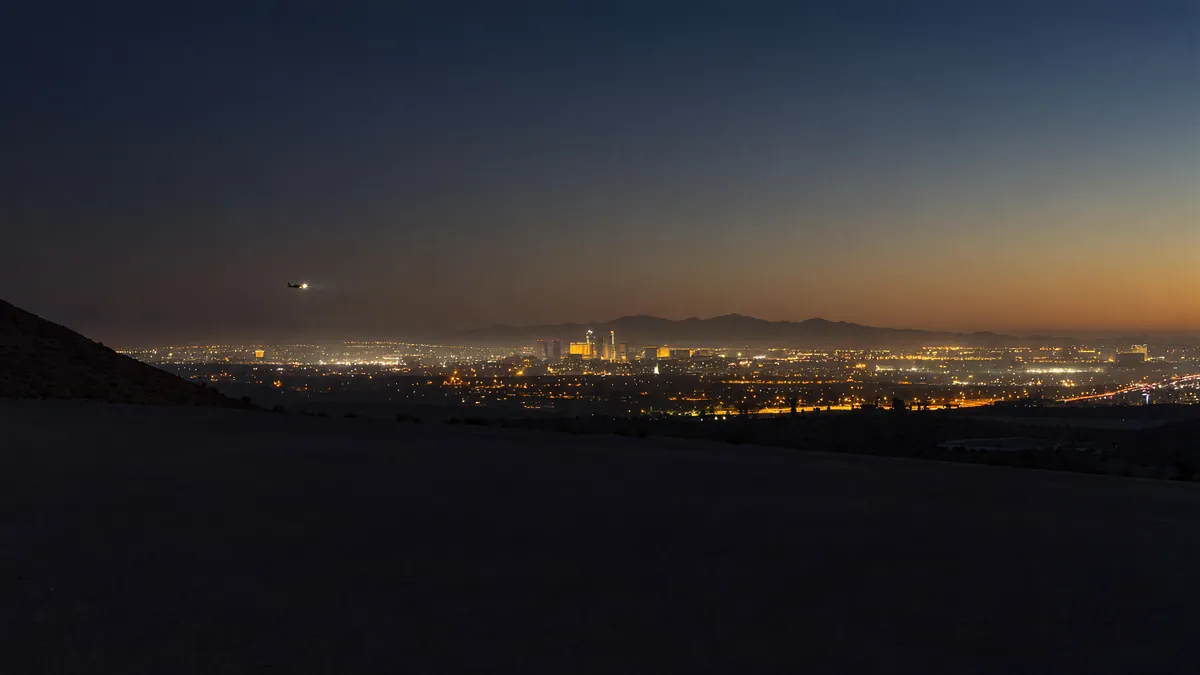 The Las Vegas Valley at dusk from a desert foothill: city lights across the basin, the Strip far off, a plane descending
