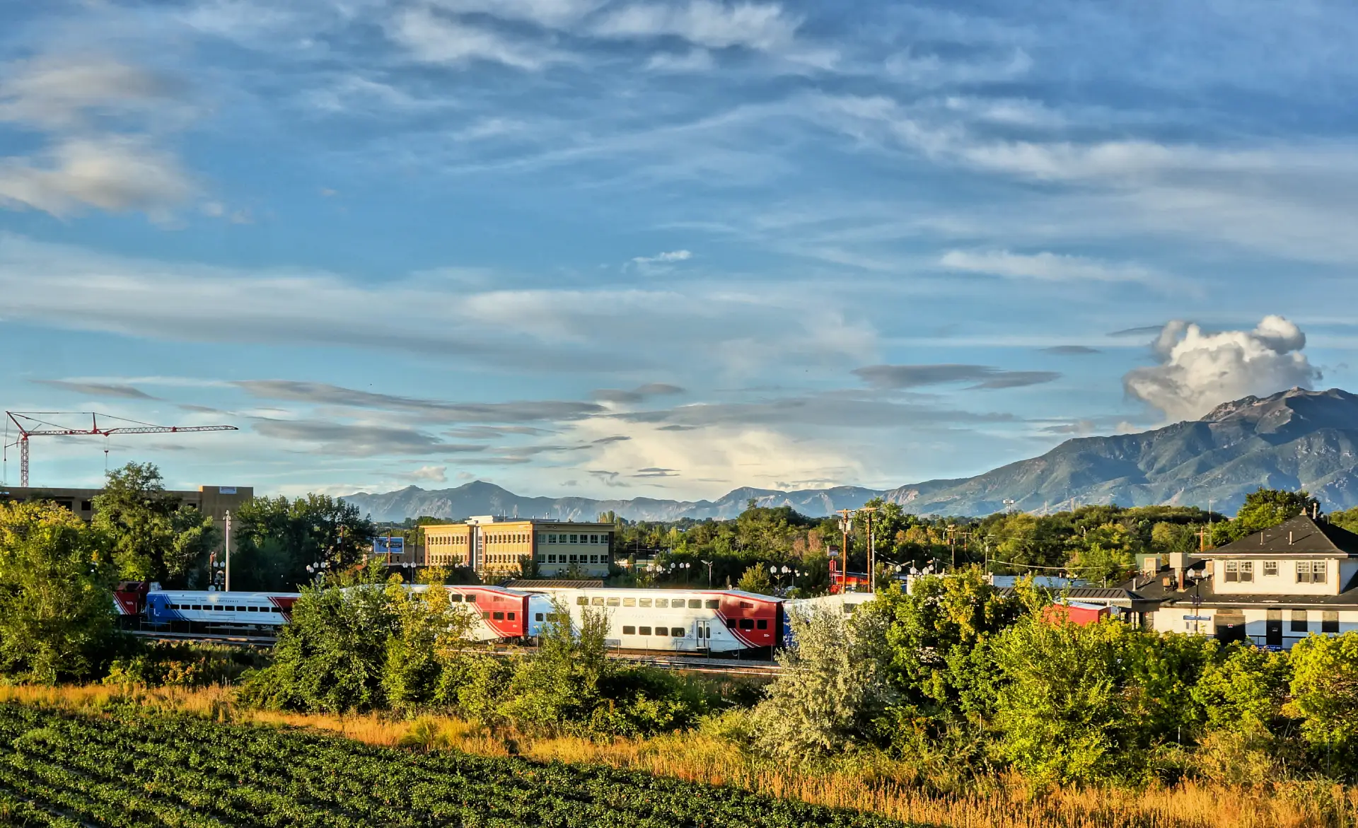 A FrontRunner commuter train running below the Wasatch Mountains in northern Utah, late-afternoon light on the fields