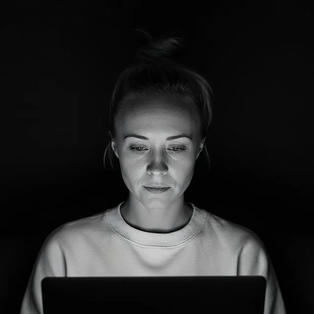 Katie, hair up and wearing a light sweater, working on a laptop in a dark room. Black and white.