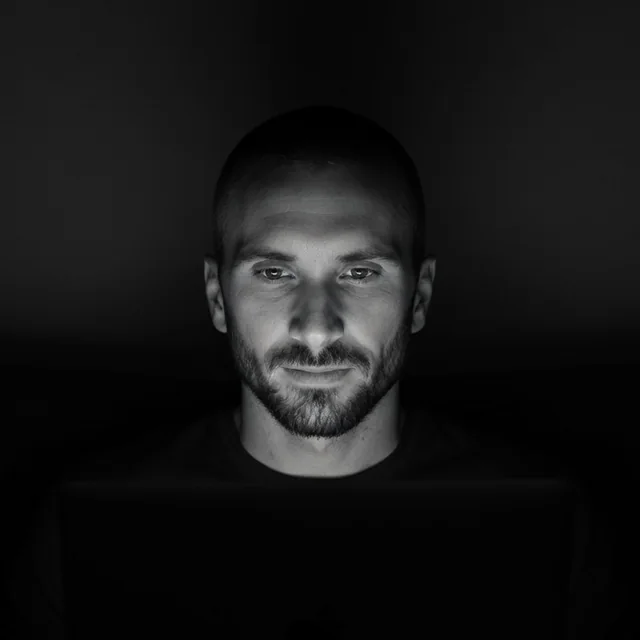 Kyle, bearded with a close-cropped head, lit by a laptop screen in a dark room. Black and white.