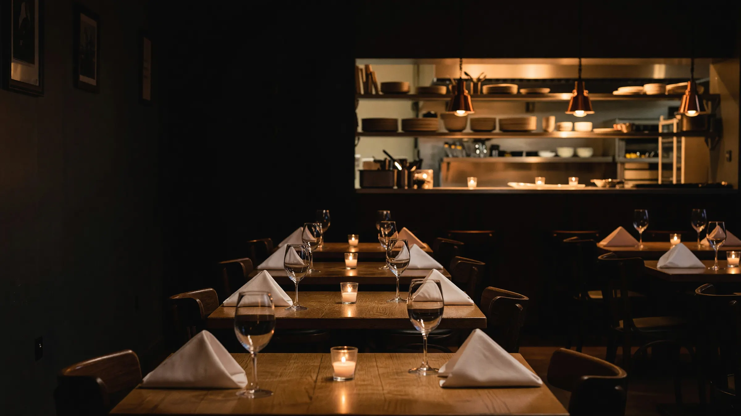 An empty restaurant dining room set for evening service, candles lit, the open kitchen pass glowing at the back
