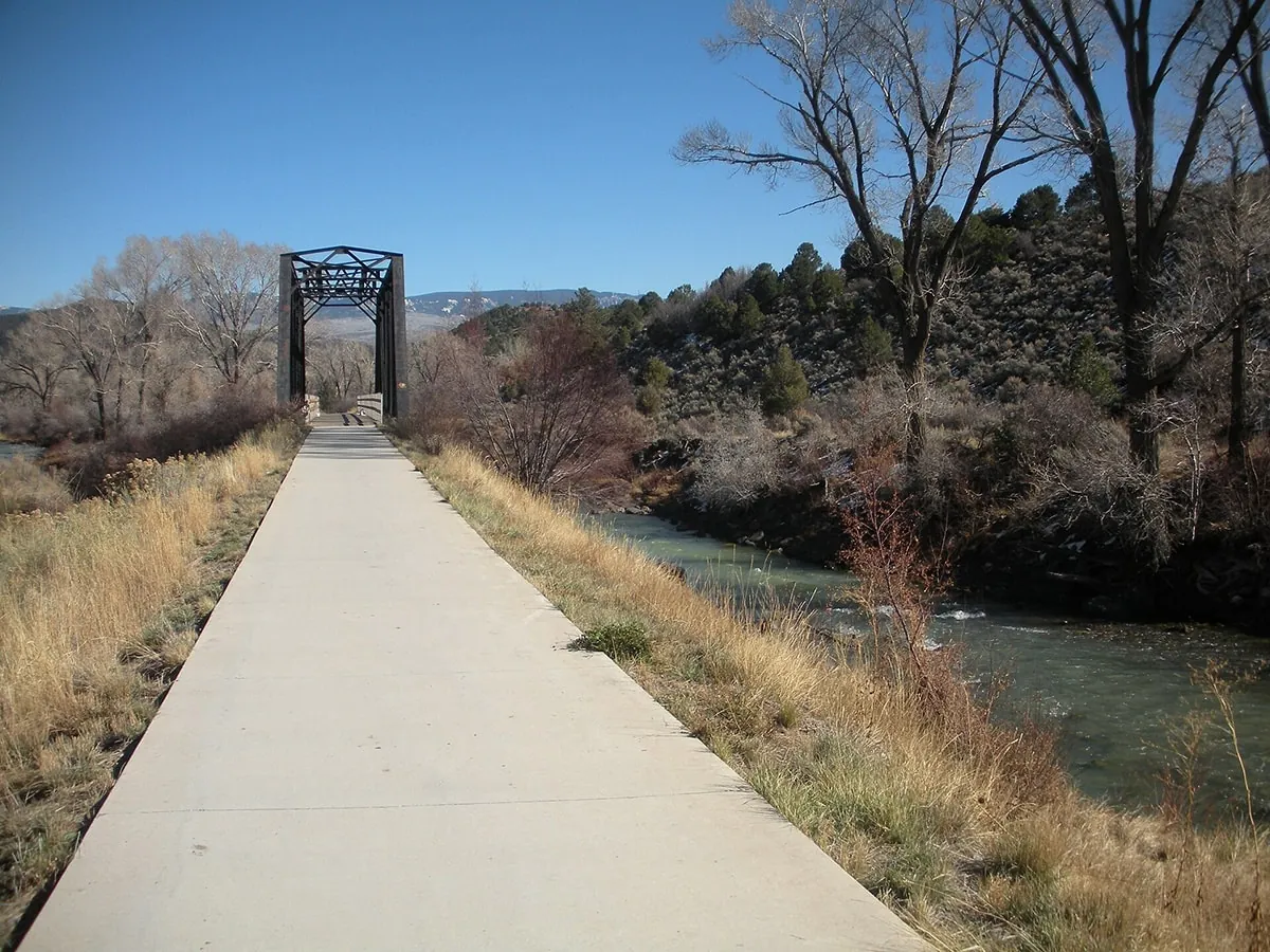 A concrete trail beside a river runs to an old black iron truss bridge, past dry gold grass and bare cottonwoods in late fall