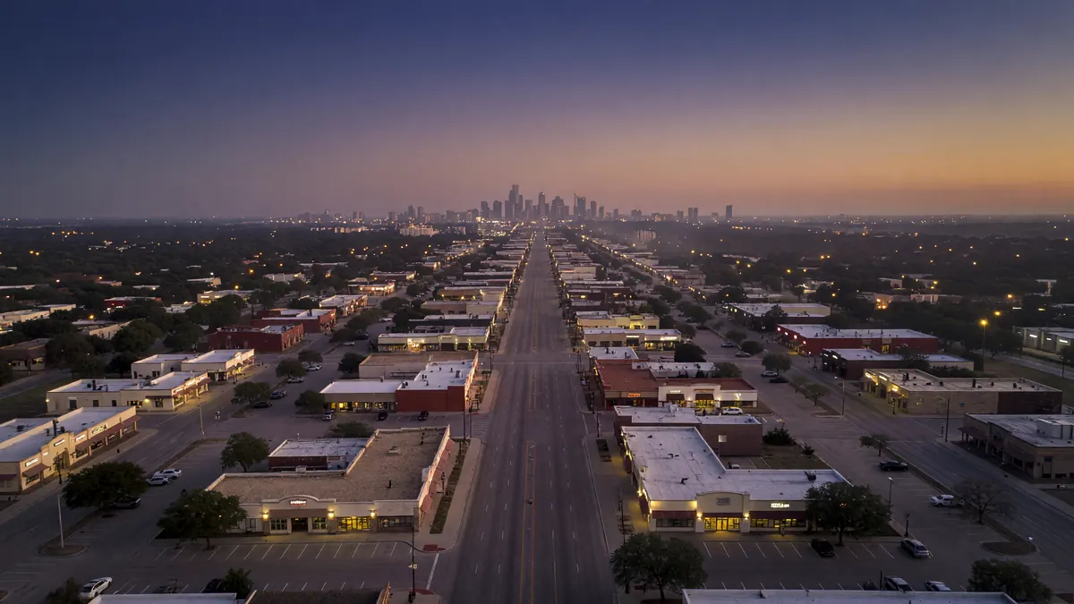 Dusk aerial of a long San Antonio commercial road lined with lit shops, the downtown skyline small on the horizon