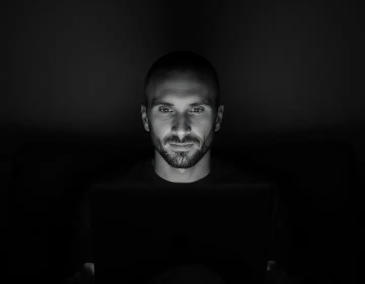 Kyle Rutten, lit by his laptop screen in a dark room, in a black and white portrait