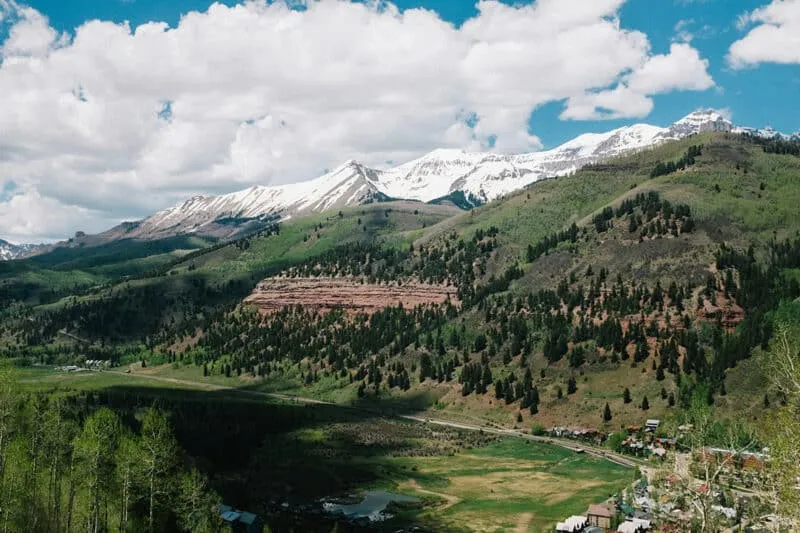 The Telluride valley in early summer: green slopes with pines and aspens, a snowy ridge under white clouds, town below.