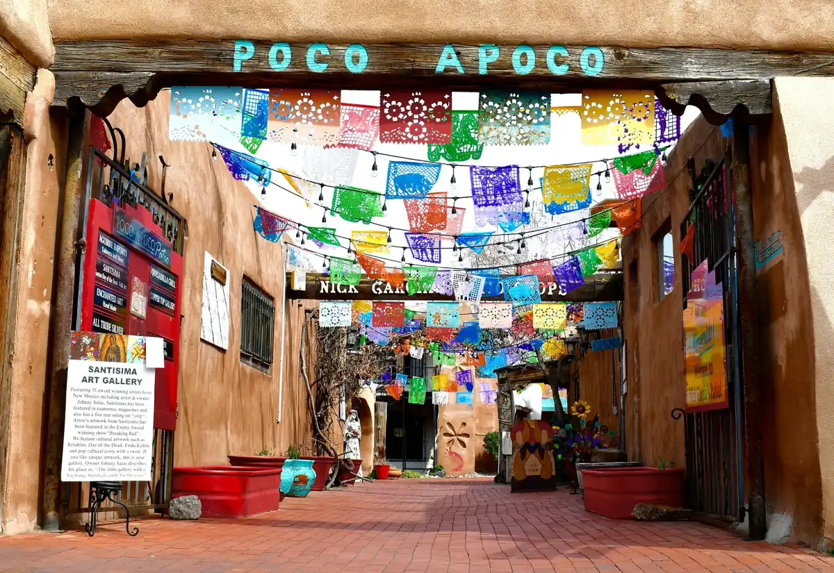 Adobe gateway into a brick courtyard in Old Town Albuquerque, with a turquoise Poco A Poco sign and papel picado overhead