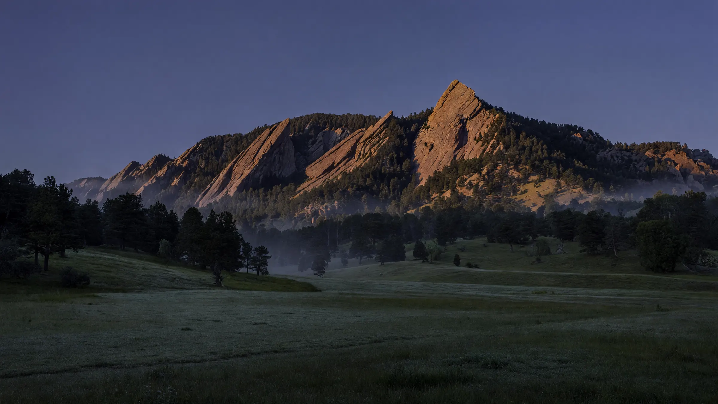 The Flatirons above Boulder at first light, sun on the sandstone slabs and haze in the pines over an open meadow