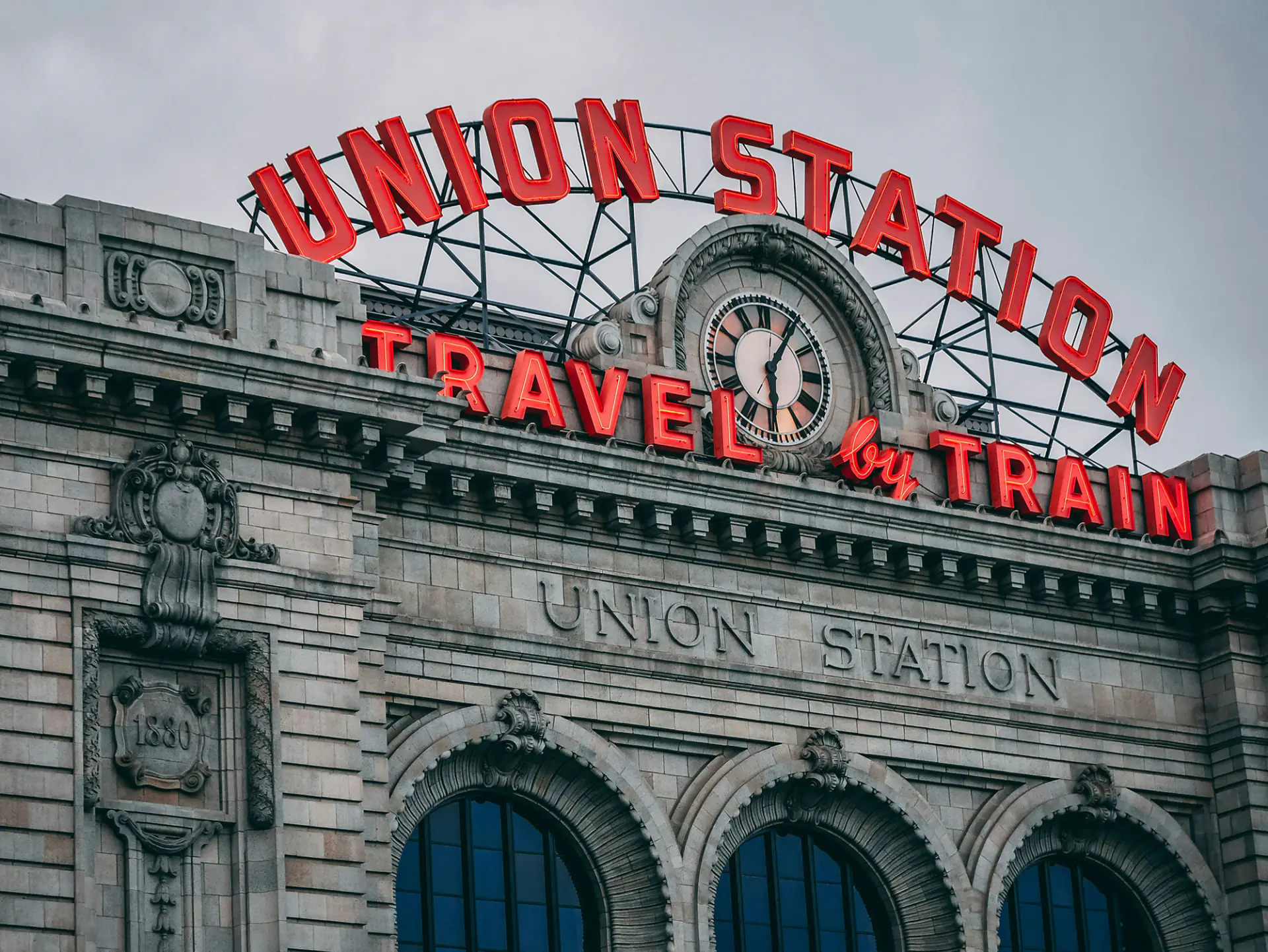 Red neon Union Station and Travel by Train signs above the clock on the stone facade of Denver Union Station