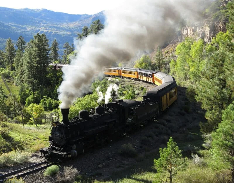 The Durango & Silverton steam train trailing smoke as its black locomotive pulls yellow cars around a bend through the pines