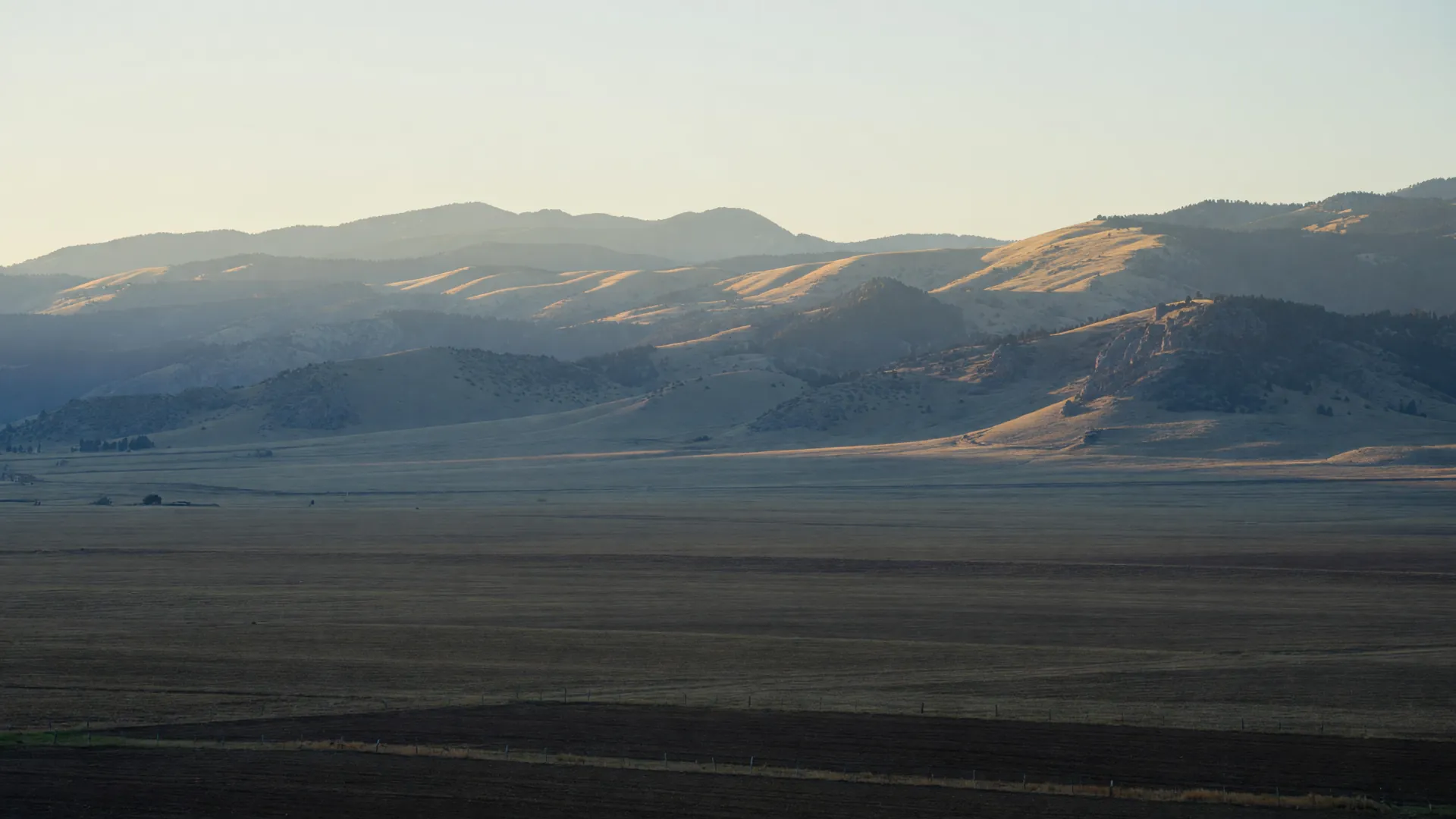 Colorado Front Range foothills at first light, layered ridgelines in soft haze above a plowed farm field