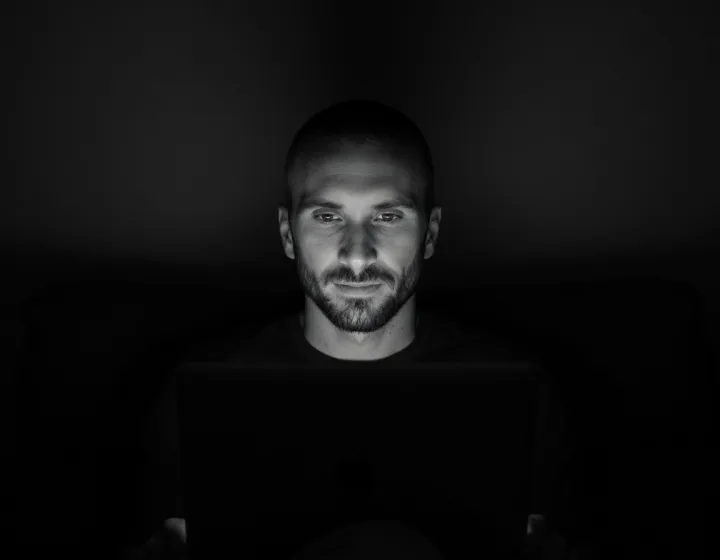 Kyle Rutten, who builds the sites, lit by the glow of his laptop in a dark room, black and white.