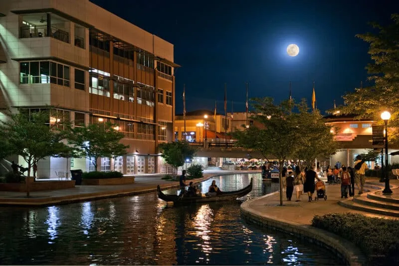 A full moon over Pueblo's Historic Arkansas Riverwalk at night, a gondola on the lit channel and people on the walkway